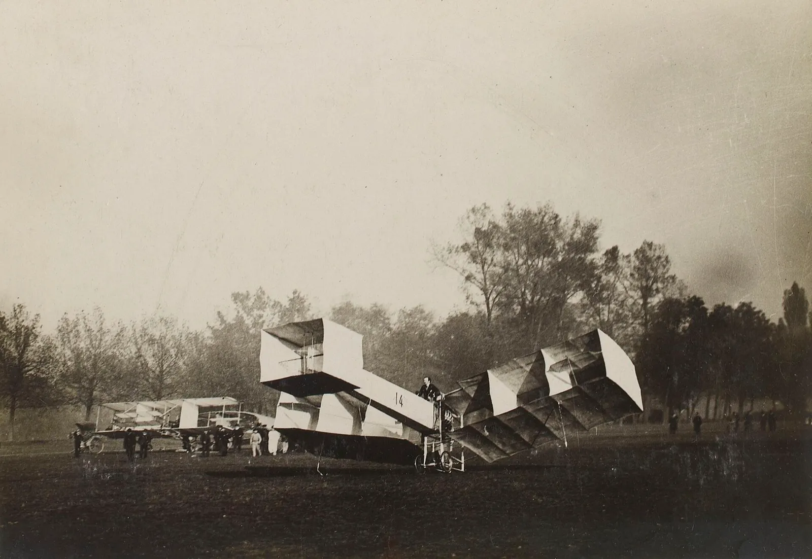 Fotografia em preto e branco do 14-Bis, aeroplano de Santos Dumont, em um gramado com árvores ao fundo e pessoas ao longe.