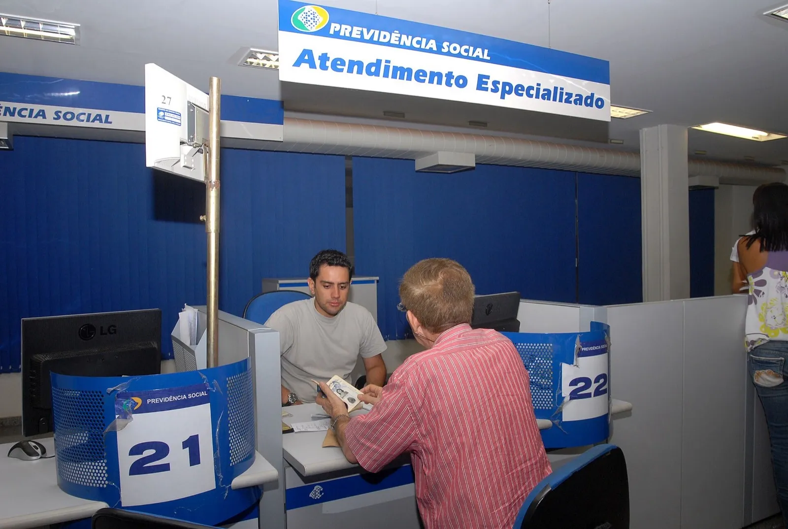 Um homem de camisa listrada é atendido por um servidor no guichê 21 de uma agência da Previdência Social, sob a placa Atendimento Especializado.