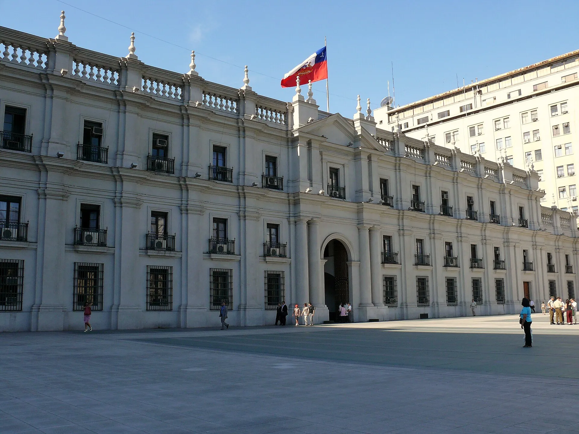 Fachada branca do Palácio de La Moneda, em Santiago, com a bandeira do Chile no alto e pessoas caminhando na praça