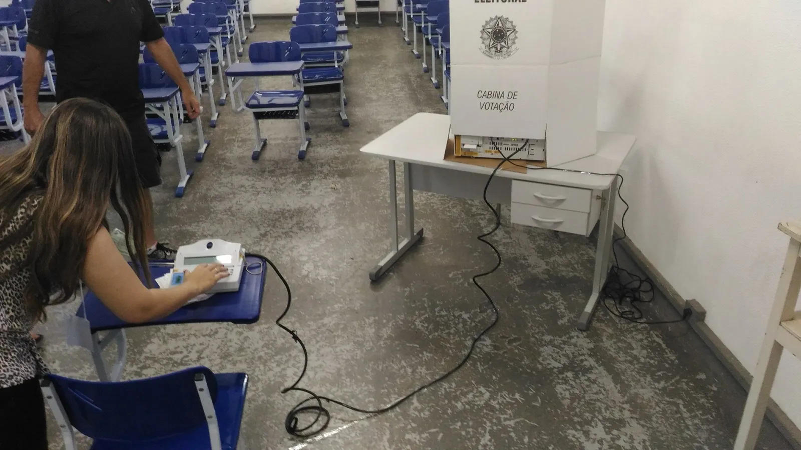 Mulher opera o terminal de uma urna eletrônica em sala de aula, ao lado de uma cabine de votação branca