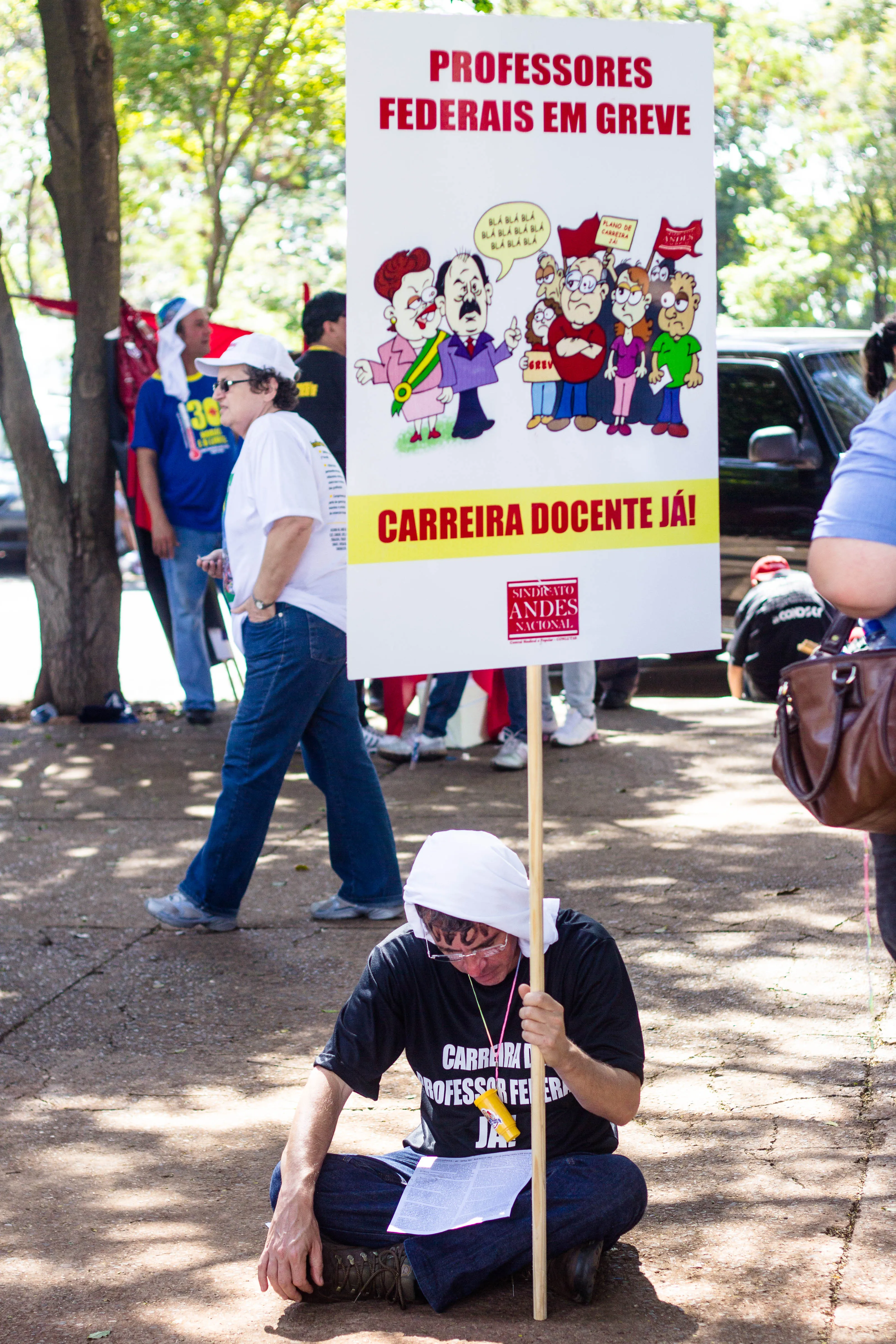 Homem sentado sob árvores segura cartaz com o título Professores federais em greve, em manifestação de servidores federais