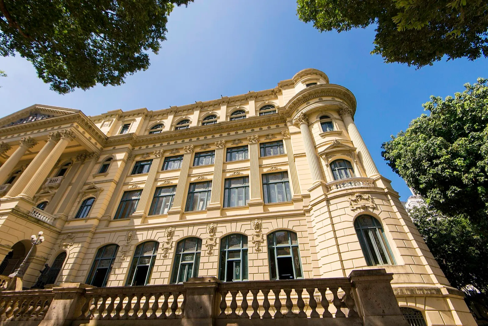 Fachada amarela e neoclássica da Biblioteca Nacional, no centro do Rio de Janeiro, com janelas altas, colunas e árvores em primeiro plano sob céu azul.