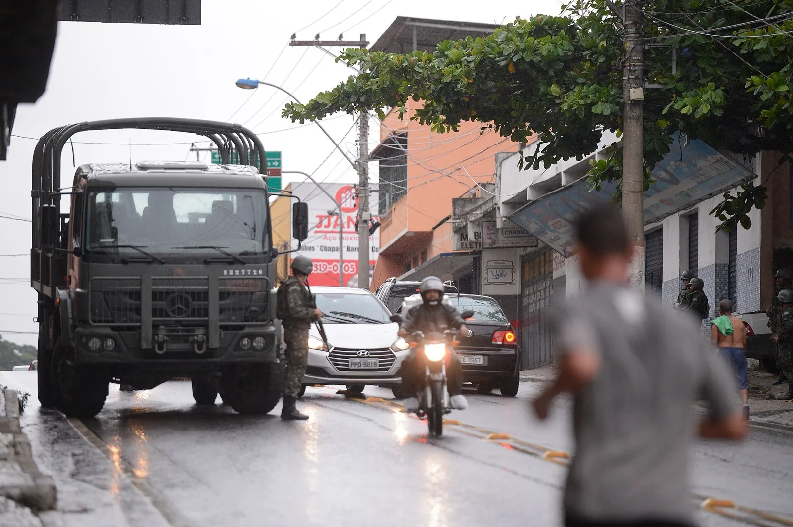 Caminhão militar verde e soldado do Exército em rua molhada de Vila Velha, com carros e uma motocicleta no trânsito