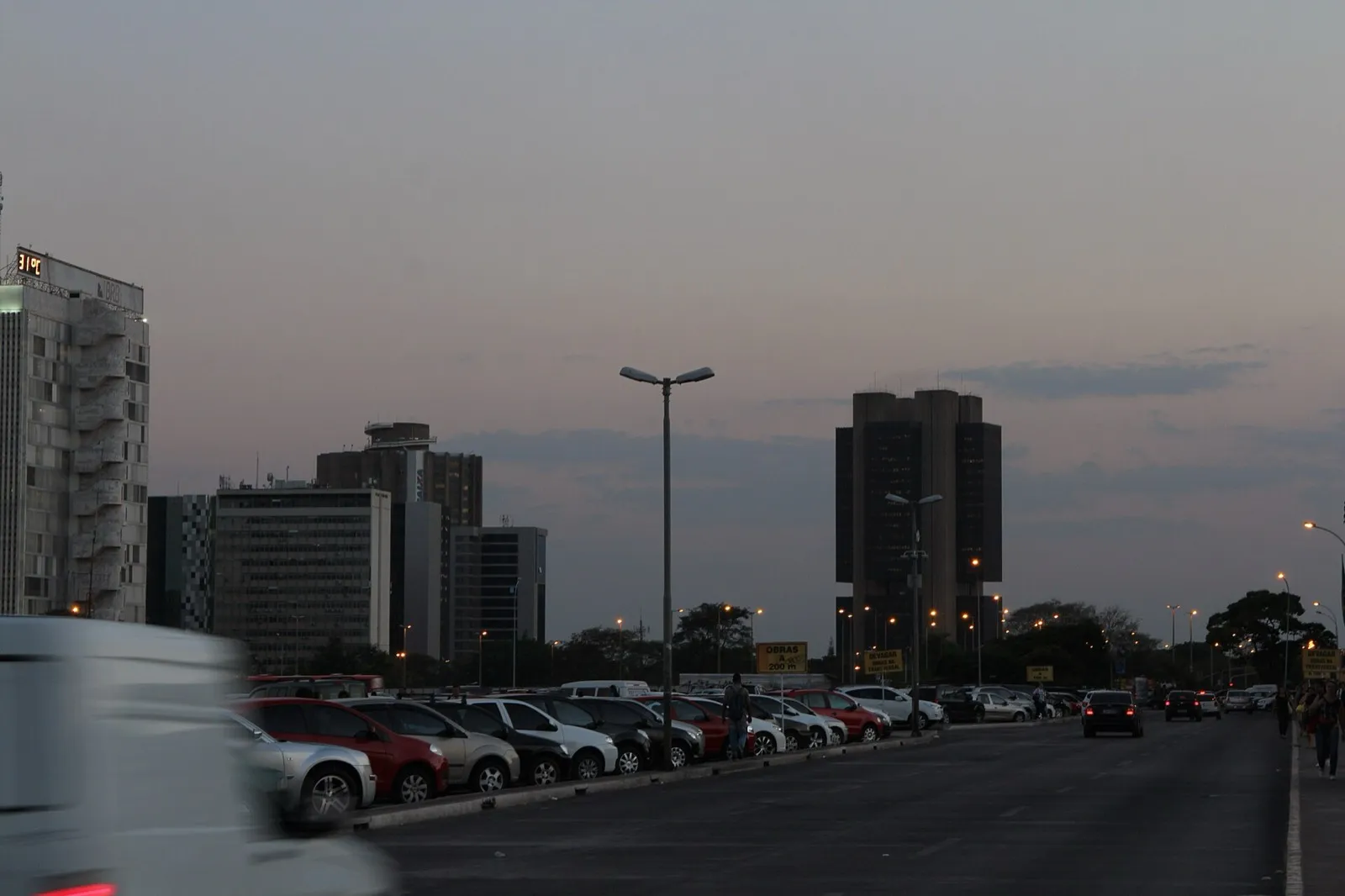 Prédio escuro da sede do Banco Central, em Brasília, visto ao anoitecer, com carros estacionados e uma avenida em primeiro plano.