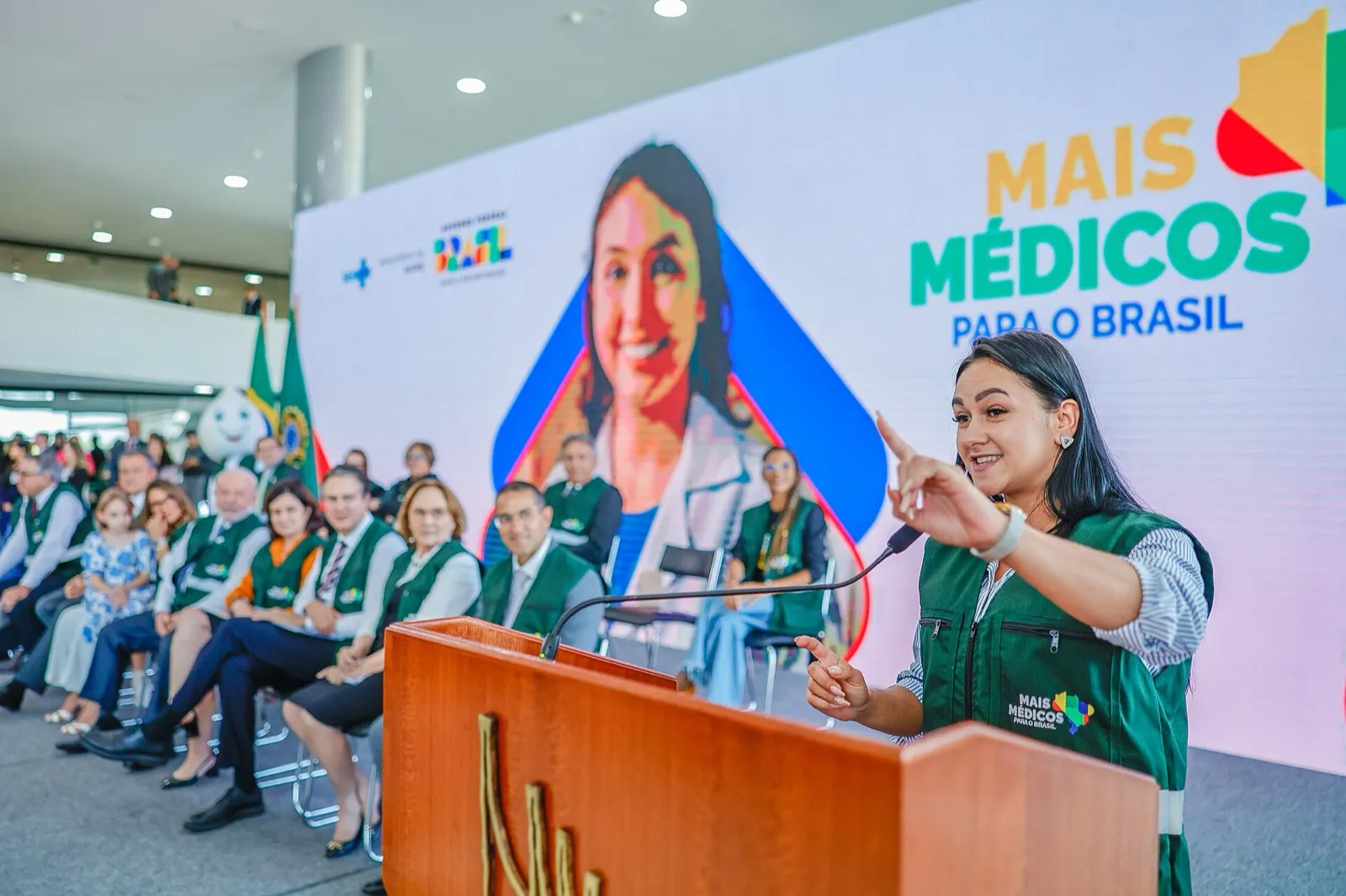 Mulher de colete verde fala ao microfone em púlpito de madeira, diante do painel Mais Médicos para o Brasil.