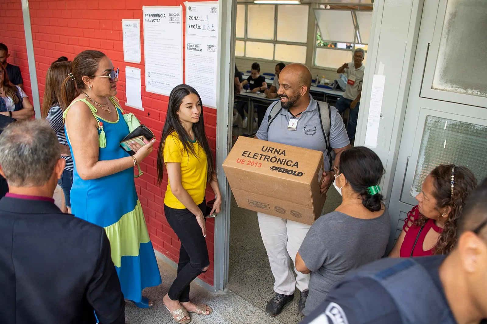 Funcionário da Justiça Eleitoral carrega uma caixa de urna eletrônica na entrada de uma seção, onde eleitores aguardam.
