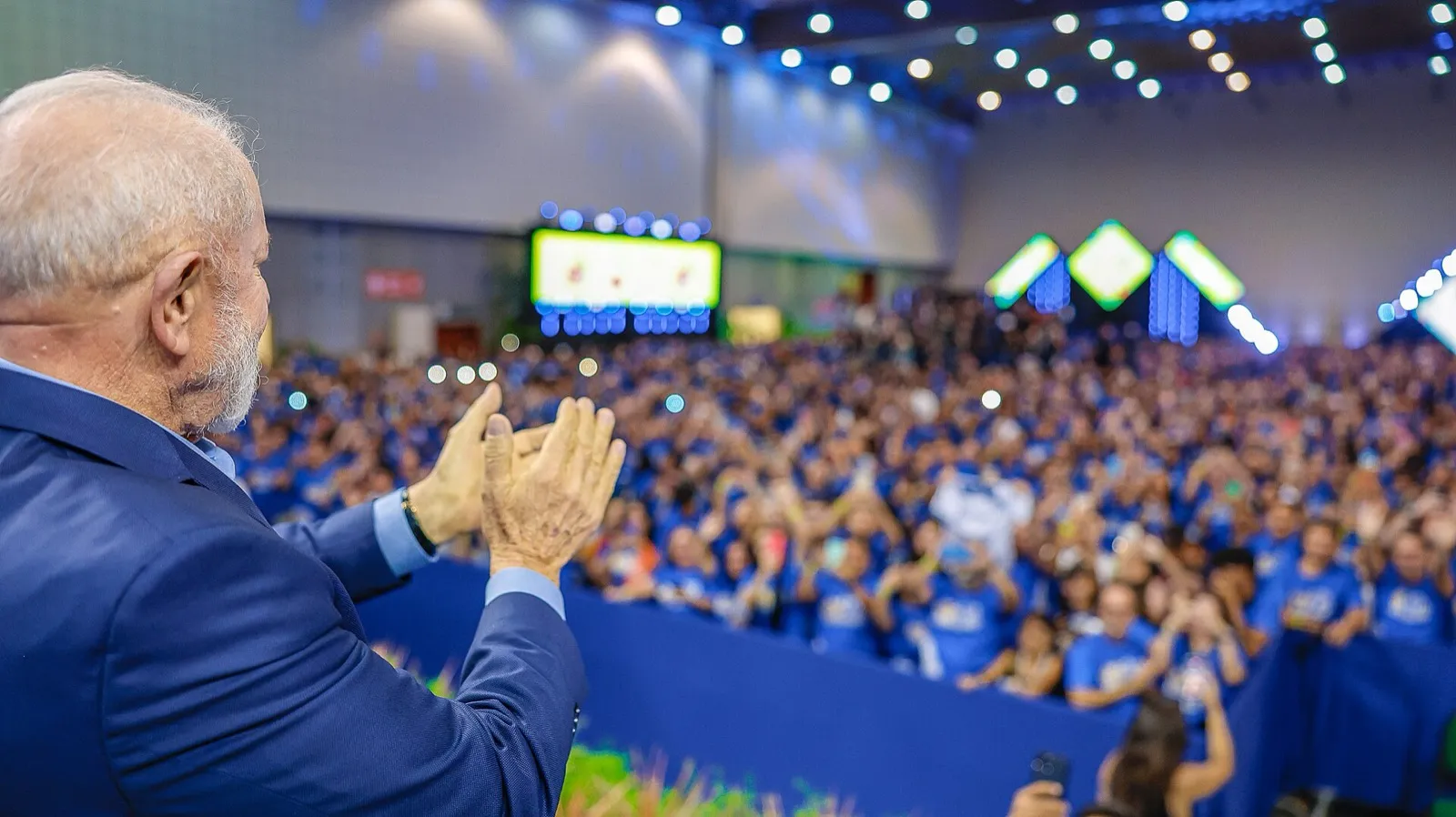 O presidente Lula aplaude, de costas para a câmera, uma plateia numerosa de camisetas azuis em um ginásio.