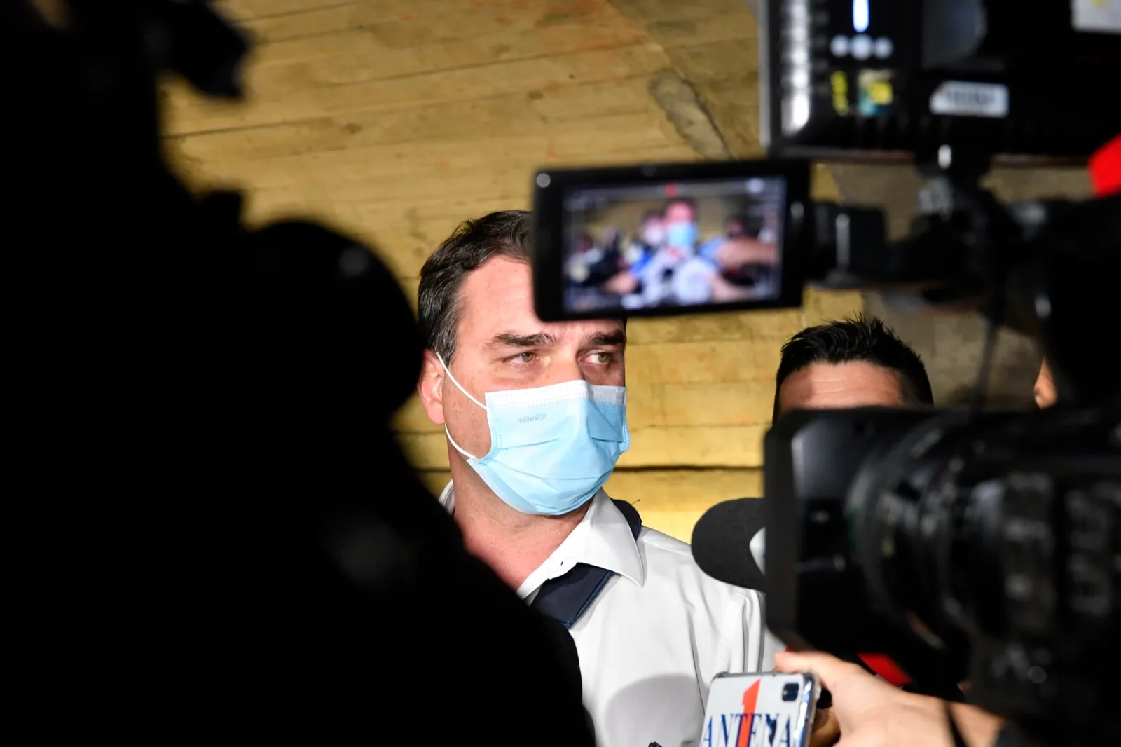 Flávio Bolsonaro, de máscara cirúrgica azul e camisa branca, fala a jornalistas entre câmeras de televisão e microfones.