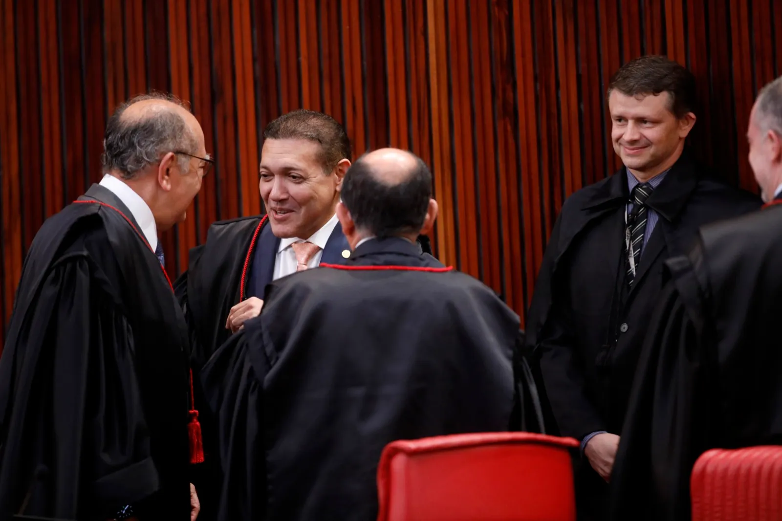 Ministros de toga conversam de pé no plenário do Tribunal Superior Eleitoral, diante de painel de madeira e cadeiras vermelhas.