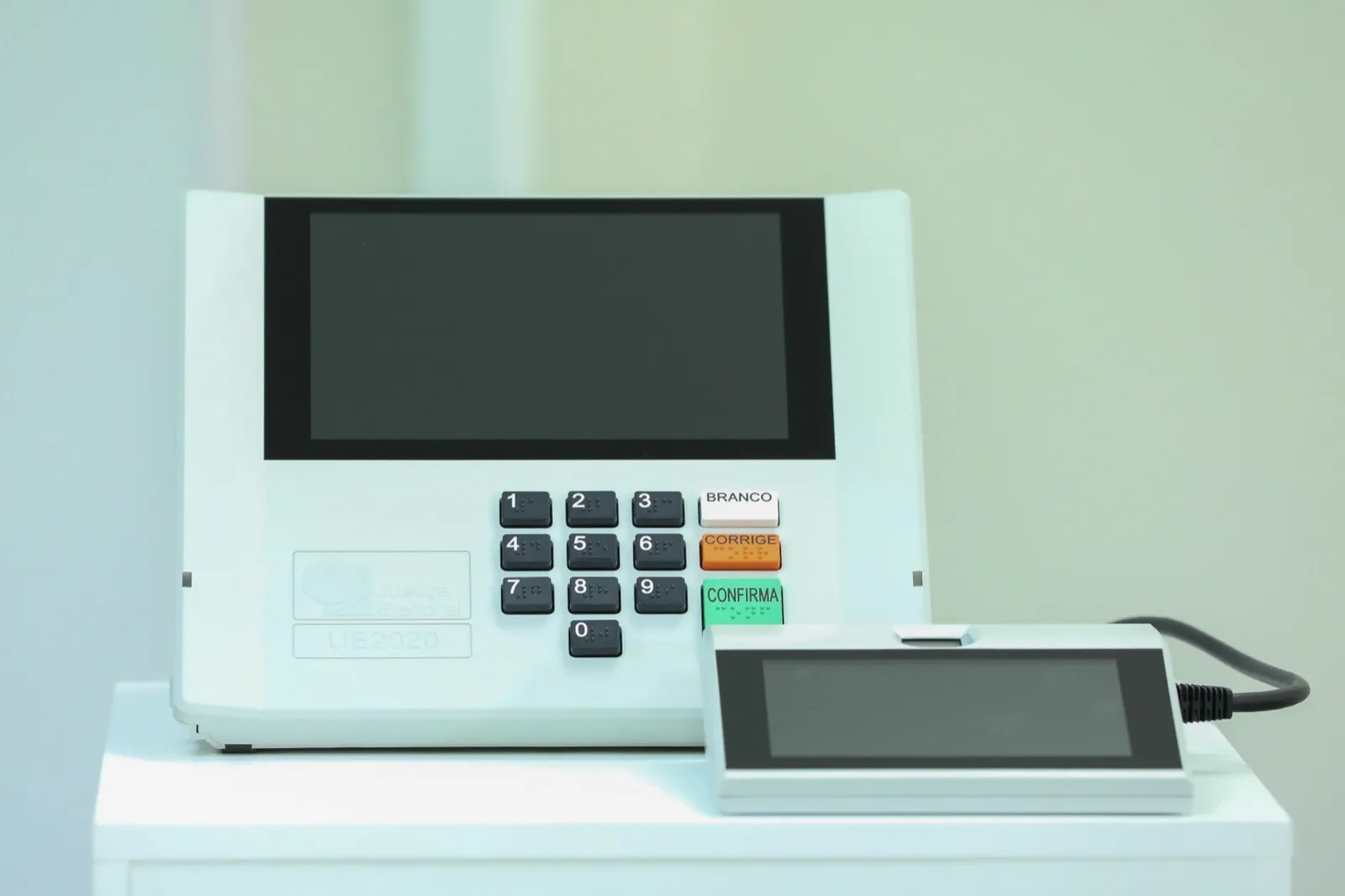 Urna eletrônica brasileira modelo UE2020, branca, com teclado numérico e as teclas Branco, Corrige e Confirma, ao lado do terminal do mesário.