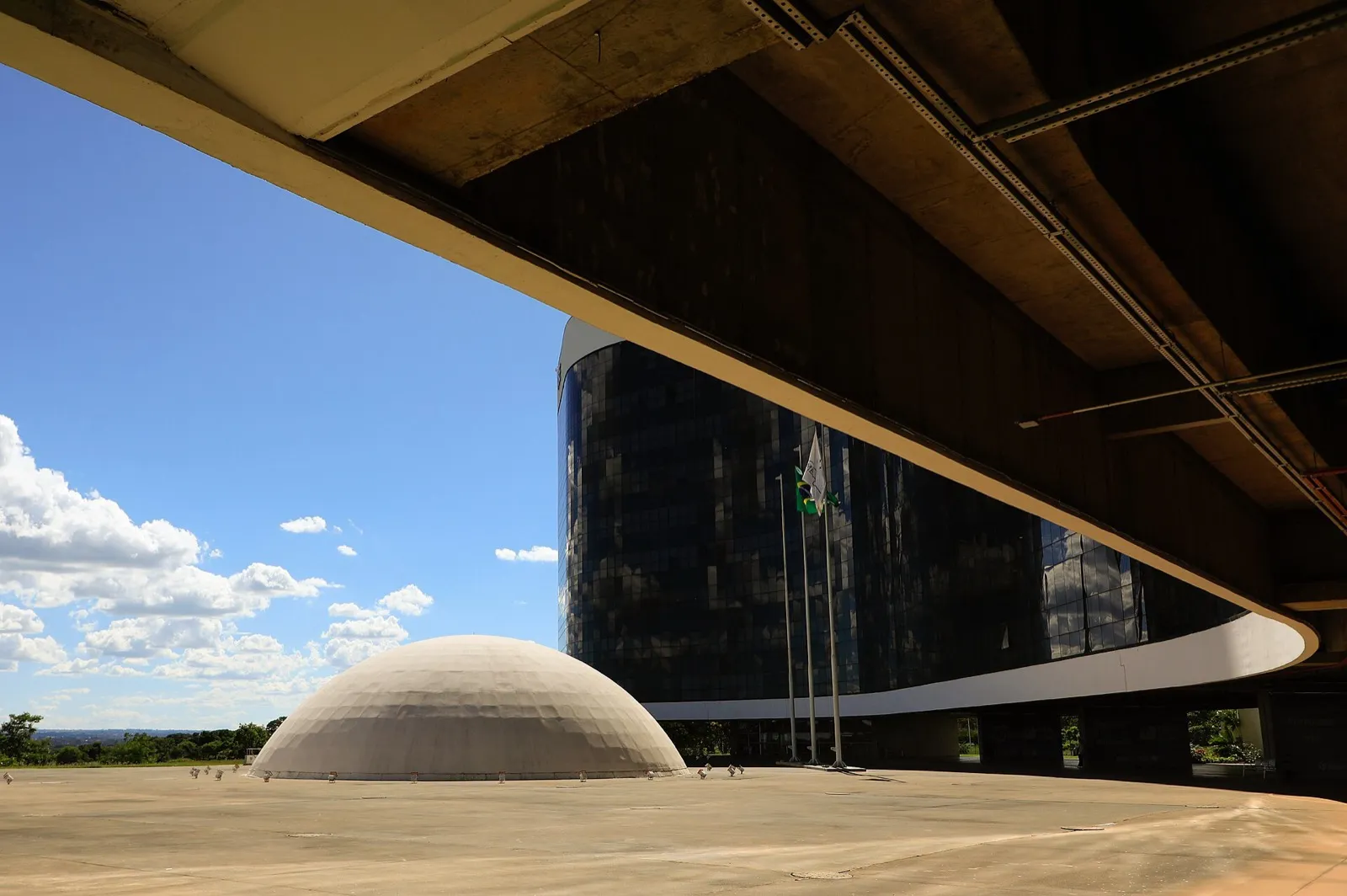 Prédio-sede do Tribunal Superior Eleitoral, em Brasília, com fachada de vidro escuro, curva branca e cúpula clara em primeiro plano, sob céu azul