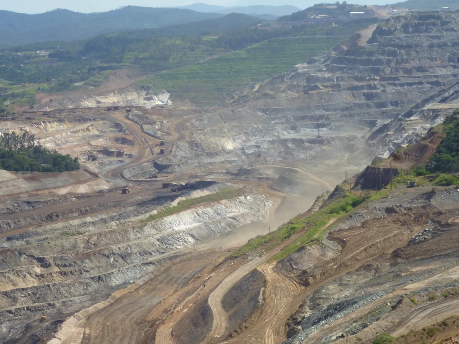 Vista do alto de uma grande mina a céu aberto, com bancadas escalonadas de terra cinza e marrom, estradas internas e morros verdes ao fundo.