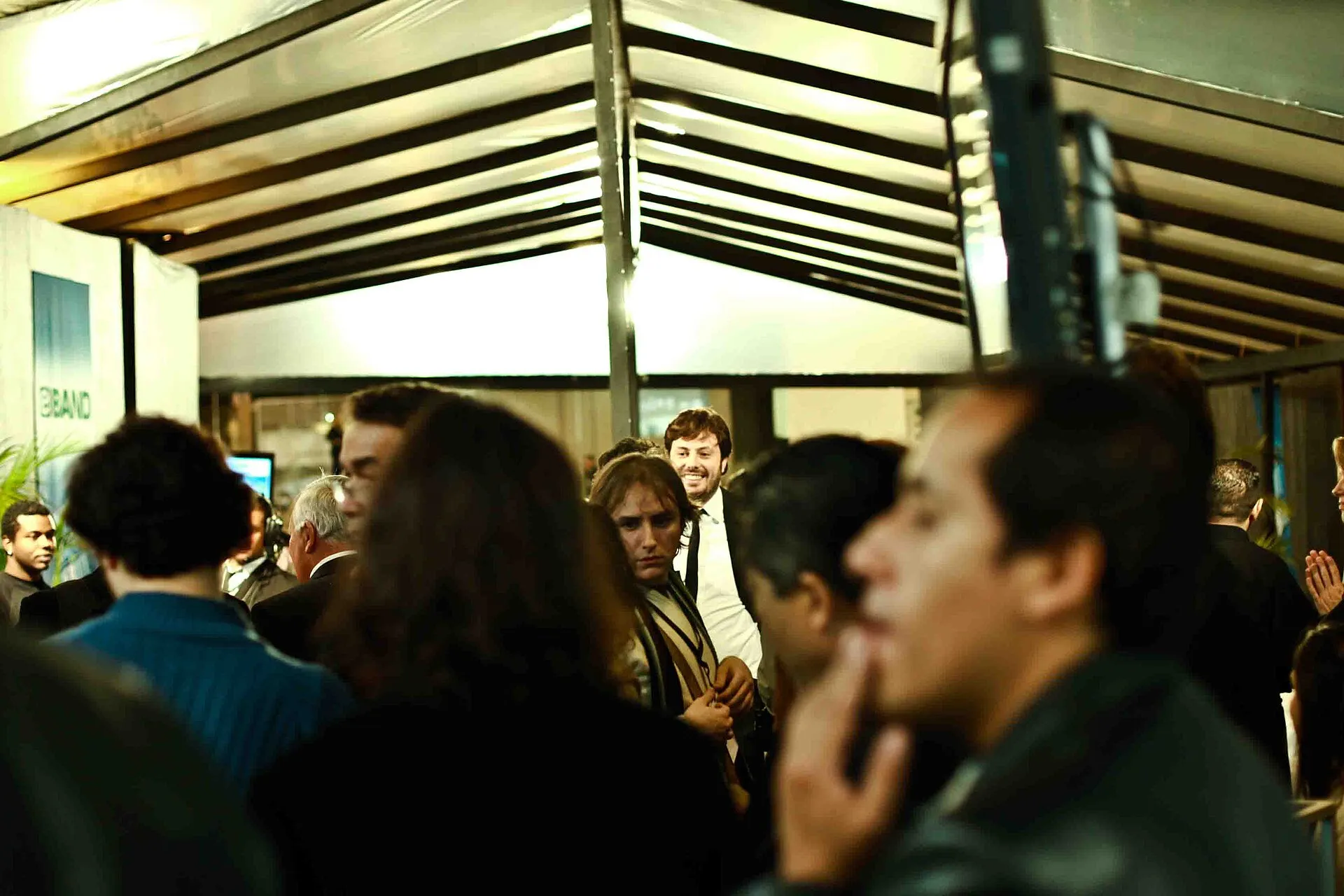 Pessoas aglomeradas sob uma cobertura metálica, em área de bastidores de um debate eleitoral; ao fundo, um homem sorridente de camisa branca.
